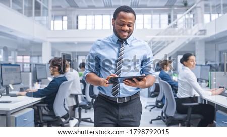 Handsome Young Black Manager in a Shirt Walking Pass His Business Colleagues with a Tablet and Supervise Their Work. Diverse and Motivated Business People Work on Computers in Modern Open Office. Royalty-Free Stock Photo #1798792015
