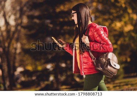 Profile side photo of positive girl tourist meet friend go walk autumn park use smartphone want find place hold bag backpack wear red coat jacket outerwear