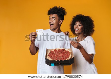 Image of delighted african american couple using credit card and cellphone while holding pizza isolated over yellow wall