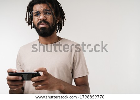 Photo of nervous emotional african guy playing games by mobile phone isolated over grey wall background in eyeglasses