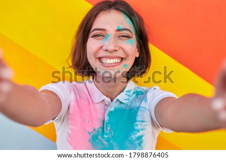 Carefree young woman with stains of paint on face and t shirt smiling and looking at camera while taking selfie during festival against colorful wall