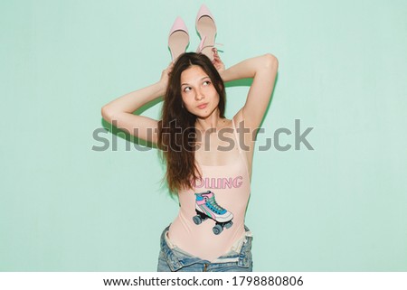 young beautiful hipster woman posing against blue wall, denim vintage style, pink swimsuit, blue jeans, holding pair of trendy shoes in hands