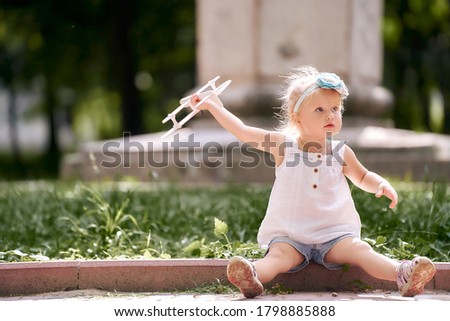 A cute little girl sits on the side of the road and holds a children's toy airplane in her outstretched hand.