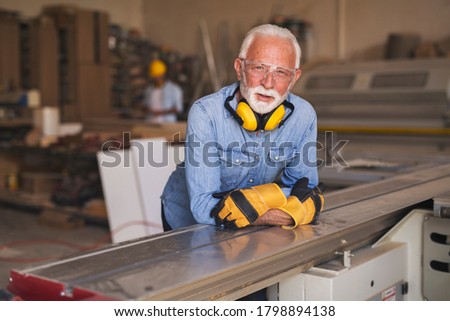 Serious senior joiner leaned on a workbench, wearing work gloves, ear protection and a protective goggles, looking at the camera