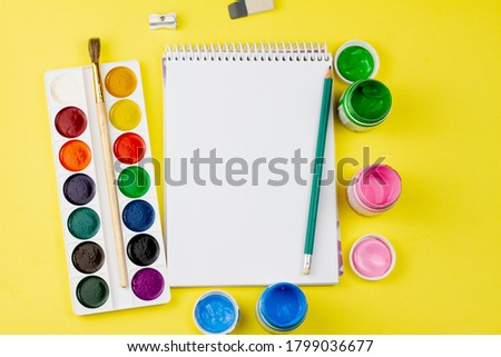 Blank notebook with colored paint and other supplies on a yellow background. View from above. Place for your text.