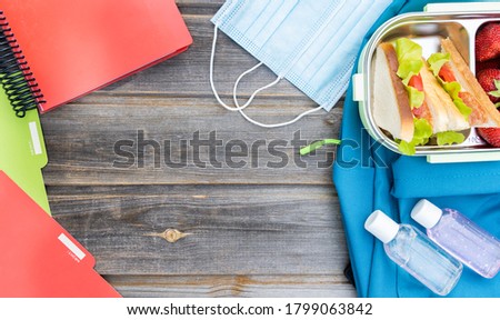 Face mask, lunchbox with sandwiches, strawberries, bottles of sanitizer. Blue backpack with notebooks, pens, box on wooden background. Back to school. Lunch with safety precautions after coronavirus.