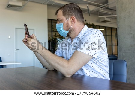 Middle aged man in a protective mask uses a smartphone while sitting in the office.