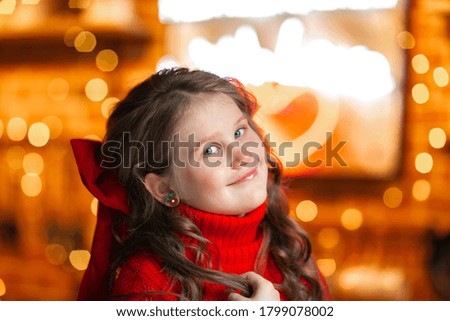 Portrait of pretty young girl in red winter sweater with curly hairdressing with red bow smiling against Christmas lights.