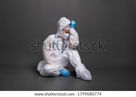 Tired female doctor in a protective suit and mask sitting on the floor on a gray background. Emergency medicine and ambulance during COVID-19