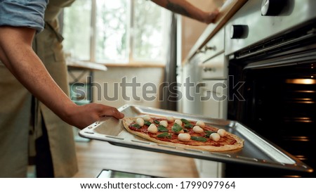 Cropped shot of man, professional cook making pizza at home. Man in apron putting raw pizza in modern oven for baking. Hobby, lifestyle. Selective focus. Side view. Web Banner