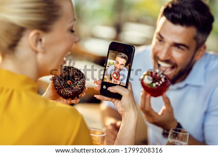 Close-up of couple having fun while eating donuts in a cafe and taking picture with mobile phone. Focus is on man on phone's screen. 
