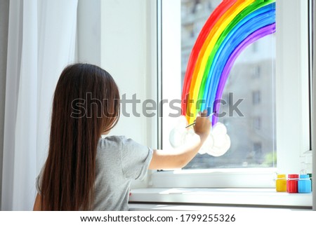 Little girl drawing rainbow on window indoors, back view. Stay at home concept