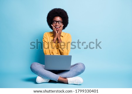 Full body size photo of black skin big volume hairstyle woman sit floor hold netbook astonished smiling made successful app wear specs jeans yellow shirt isolated blue color background