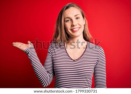 Young beautiful blonde woman wearing casual striped t-shirt over isolated red background smiling cheerful presenting and pointing with palm of hand looking at the camera.