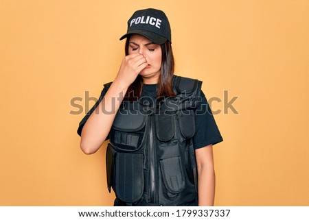 Young beautiful brunette policewoman wearing police uniform bulletproof and cap smelling something stinky and disgusting, intolerable smell, holding breath with fingers on nose. Bad smell