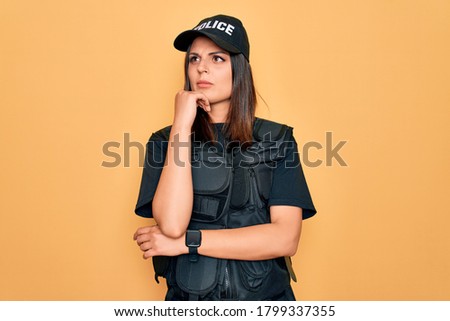 Young beautiful brunette policewoman wearing police uniform bulletproof and cap with hand on chin thinking about question, pensive expression. Smiling with thoughtful face. Doubt concept.