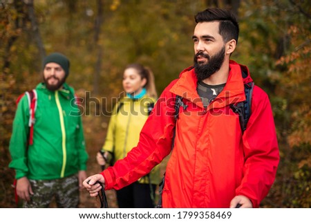 Young bearded male backpacker in bright red outerwear looking away with interest while standing with friends in autumn forest and exploring nature