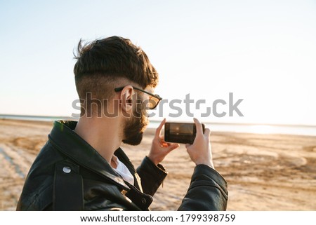 Back view of an stylish young bearded man taking picture on mobile phone while standing at the sunny beach
