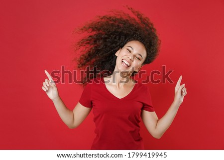 Funny young african american woman 20s wearing basic casual burgundy t-shirt isolated on red background, studio portrait. Having fun dancing with flying fluttering hair, pointing index fingers up