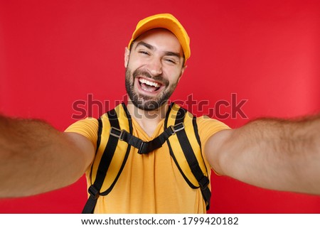 Close up delivery man 20 in yellow cap t-shirt uniform thermal bag backpack doing selfie isolated on red background studio Guy male employee work as courier Service coronavirus covid-19 virus concept