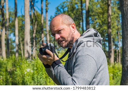 Adult man in hoodie holding mirror less camera, preparing to shoot photos in a forest in summer day, half body portrait
