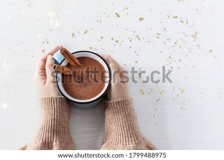 Hot chocolate with cinnamon spice in enamel mug in female hands on white wooden background top view. Hot cozy drink for autumn or winter season. 