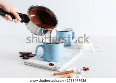 Person hand pouring hot chocolate in a pot into blue enamel cups over white background. Hot cozy drink for autumn or winter season.