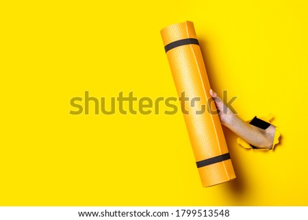 Female hand holding an orange yoga mat on a bright yellow background.