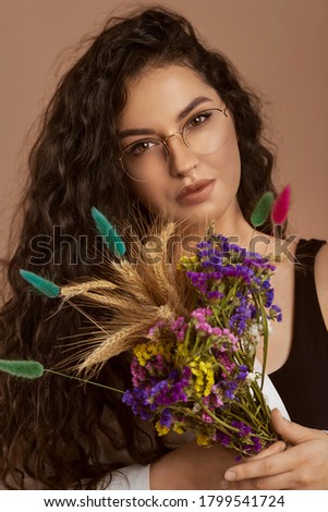 Young beautiful woman with glasses and curly hair posing with stylish dried flowers bouquet. Close-up portrait. Beauty concept. Isolated on pastel brown background.