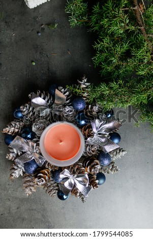 Diy Candle holder in New Year and Christmas style. Cones, blue Christmas balls, silver bows around an orange candle in a glass. Juniper branches on dark gray table