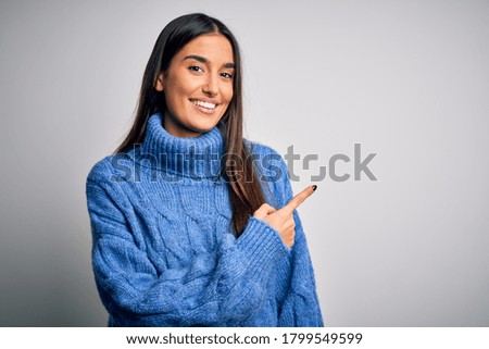 Young beautiful brunette woman wearing casual turtleneck sweater over white background cheerful with a smile of face pointing with hand and finger up to the side with happy and natural expression