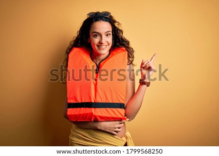 Young beautiful woman with curly hair wearing orange lifejacket over yellow background with a big smile on face, pointing with hand and finger to the side looking at the camera.
