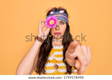 Young beautiful hippie woman with blue eyes wearing sunnglasses holding pink flower over eye pointing with finger to the camera and to you, hand sign, positive and confident gesture from the front