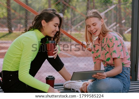 In the park handsome women have a good time together they using a electronic tablet to watch some video while sitting on the park chair