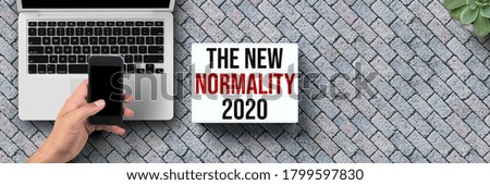lightbox with message NEW NORMALITY 2020 with laptop and hand holding smartphone on stone pavement background