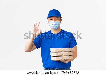Contactless delivery, covid-19 shipping, preventing virus concept. Cheerful asian male courier in blue uniform, medical mask and gloves, bring pizza to client house, ensure quality, show okay sign