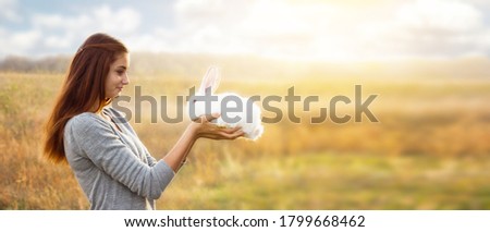 young girl with a red hair and a gray jacket is holding  small white fluffy rabbit with red eyes in front of her, smiling sweetly at him. With space for your text. On a dry steppe area. Easter concept