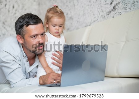 Little girl and caring father are watching cartoons on a laptop while lying on the couch at home. A man shows his little daughter photographs on a computer. Beloved daddy.