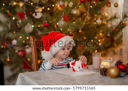 A boy in a Santa Claus hat writes a letter with wishes at the table with a drain of cocoa, on the background there is a Christmas tree decorated in the lights of a garland