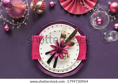 Christmas table setup with white plate, golden utensils, dark red napkin and gilded decorations. Flat lay, top view on dark purple linen textile background. Xmas lights garland.