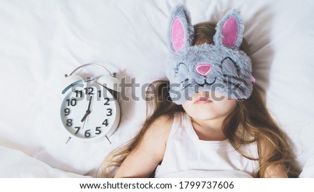 Little girl sleeping in bed under white blanket wearing grey bunny plush sleep mask with alarm clock on pillow. Early morning wake up. Putting kid to sleep.Mom's correct daily routine, rest for child. Royalty-Free Stock Photo #1799737606
