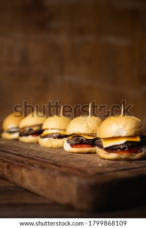 Homemade mini burguers made of ground beef, cheddar cheese, tomato and mayonnaise on a wooden table with a brown raw wooden background and space for text. 