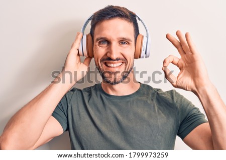 Young handsome man listening to music using headphones over isolated white background doing ok sign with fingers, smiling friendly gesturing excellent symbol