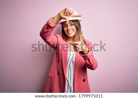 Young beautiful brunette architect woman wearing safety helmet over pink background smiling making frame with hands and fingers with happy face. Creativity and photography concept.