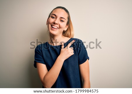 Young beautiful blonde woman with blue eyes wearing casual t-shirt over white background cheerful with a smile of face pointing with hand and finger up to the side with happy and natural expression