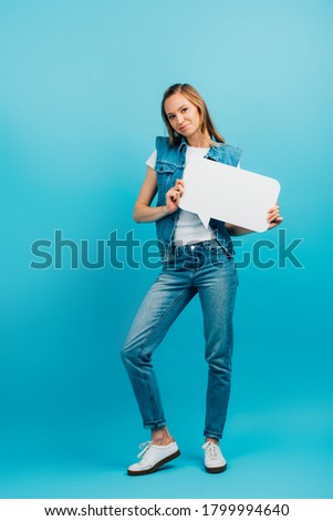 full length view of woman in denim vest and jeans holding speech bubble on blue
