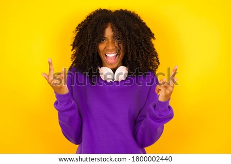 Born to rock this world. Joyful African American woman in fashionable clothes screaming out loud and showing with raised arms horns or rock gesture, expressing excitement of being on concert of band.