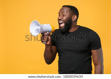 Cheerful african american man in casual black t-shirt isolated on yellow background studio portrait. People sincere emotions lifestyle concept. Mock up copy space. Scream in megaphone, looking aside Royalty-Free Stock Photo #1800039634