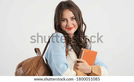 Beautiful brunette student girl with backpack and book charmingly looking in camera over white background