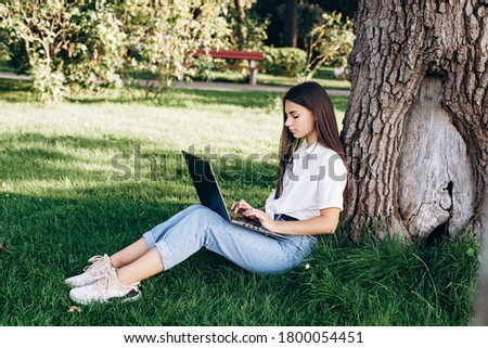 girl student with a laptop outdoors sits on the grass in the park near a tree, surfing the internet or preparing for exams. Technology, education and remote work concept. Soft selective focus.